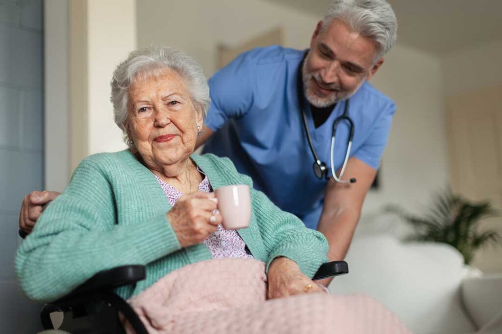 Older adult talking with a caregiver in a bright assisted living common room while holding a cup of tea